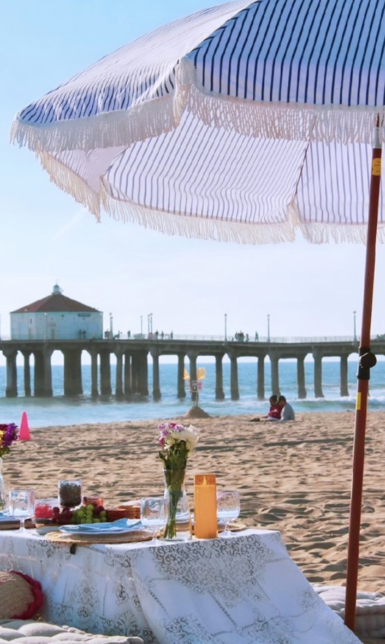 Styled low beach picnic with a fringed striped umbrella and grazing table on the sand, the Manhattan Beach pier behind