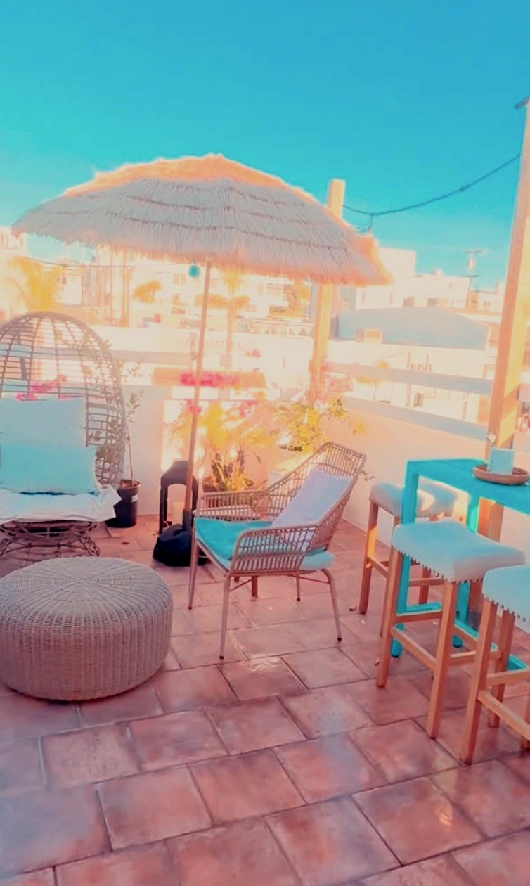 Sun-drenched rooftop patio at Tropical Refuge — thatched umbrella, rattan seating and blue bar stools on terracotta tile in Manhattan Beach